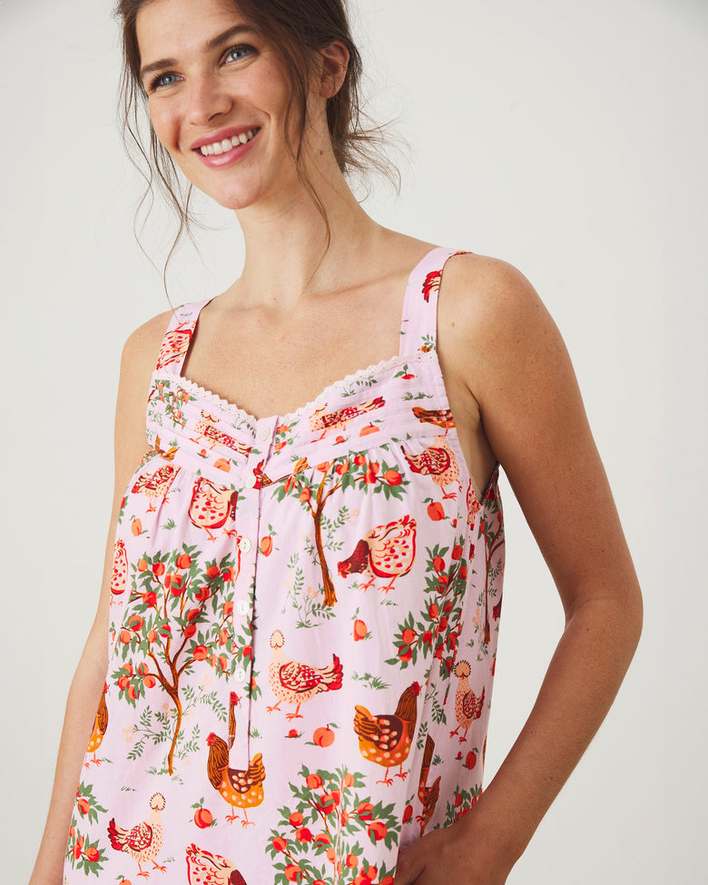 Front upper-body view of a ballet-pink nightgown showing its wide straps, scalloped neckline trim, white buttons, and hen-and-fruit print.