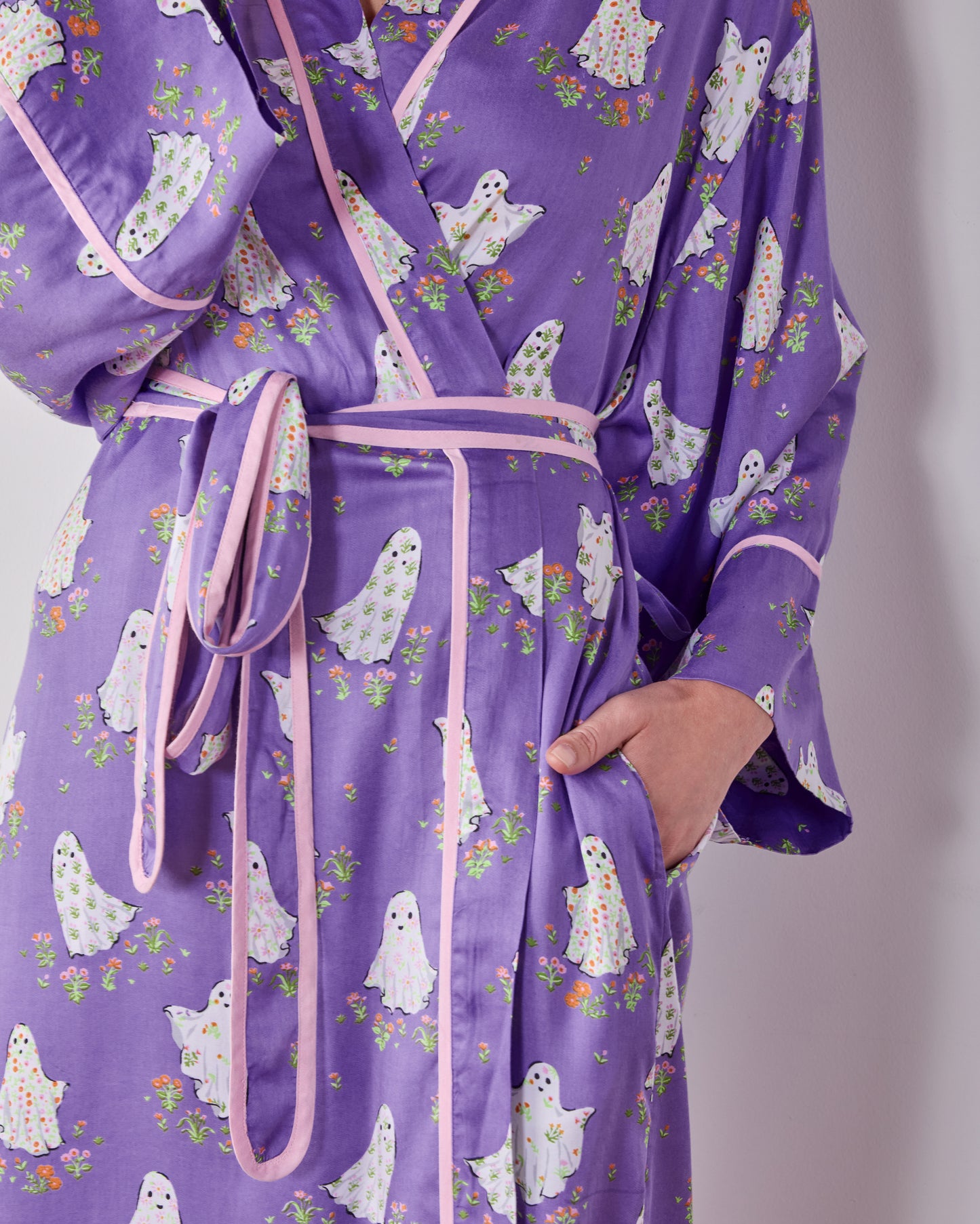 Close view of the purple ghost-and-floral print robe showing its pink-trimmed front, matching belt, and hand in a pocket.