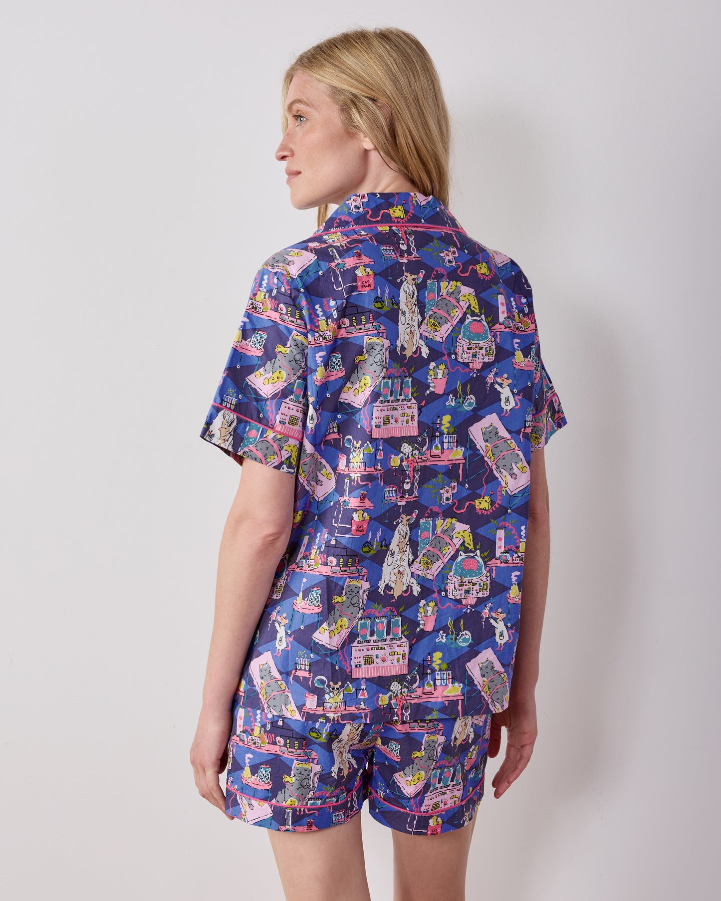 Back view of a person wearing a short-sleeved button-down pajama top and matching shorts with a colorful Frankenstein-inspired laboratory print.