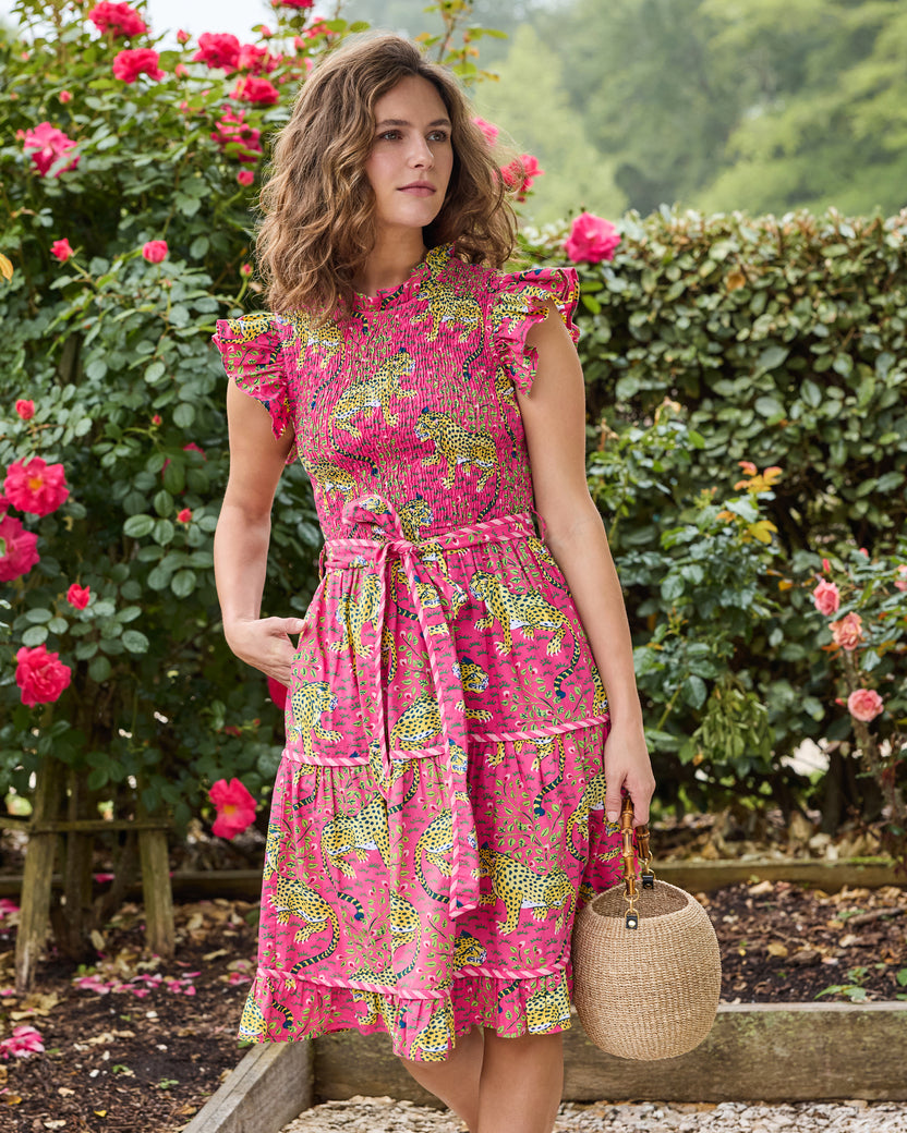 Model wearing a hot pink tiered leopard-print dress with flutter sleeves and a coordinating waist belt, holding a round woven handbag in a rose garden.