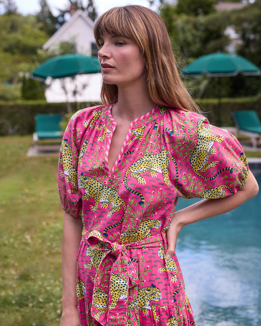 Close view of the hot pink Bagheera To and From Dress showing its leopard print, V-neckline, puff sleeves, and tied waist belt beside a swimming pool.
