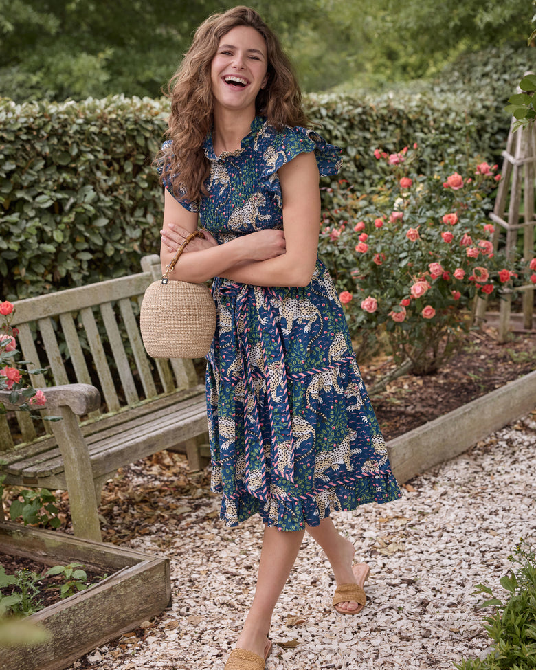 Model wearing the navy Bagheera Let's Ruffle Dress with leopard print, flutter sleeves, a smocked bodice, tiered skirt, and coordinating belt, holding a round woven handbag in a garden.