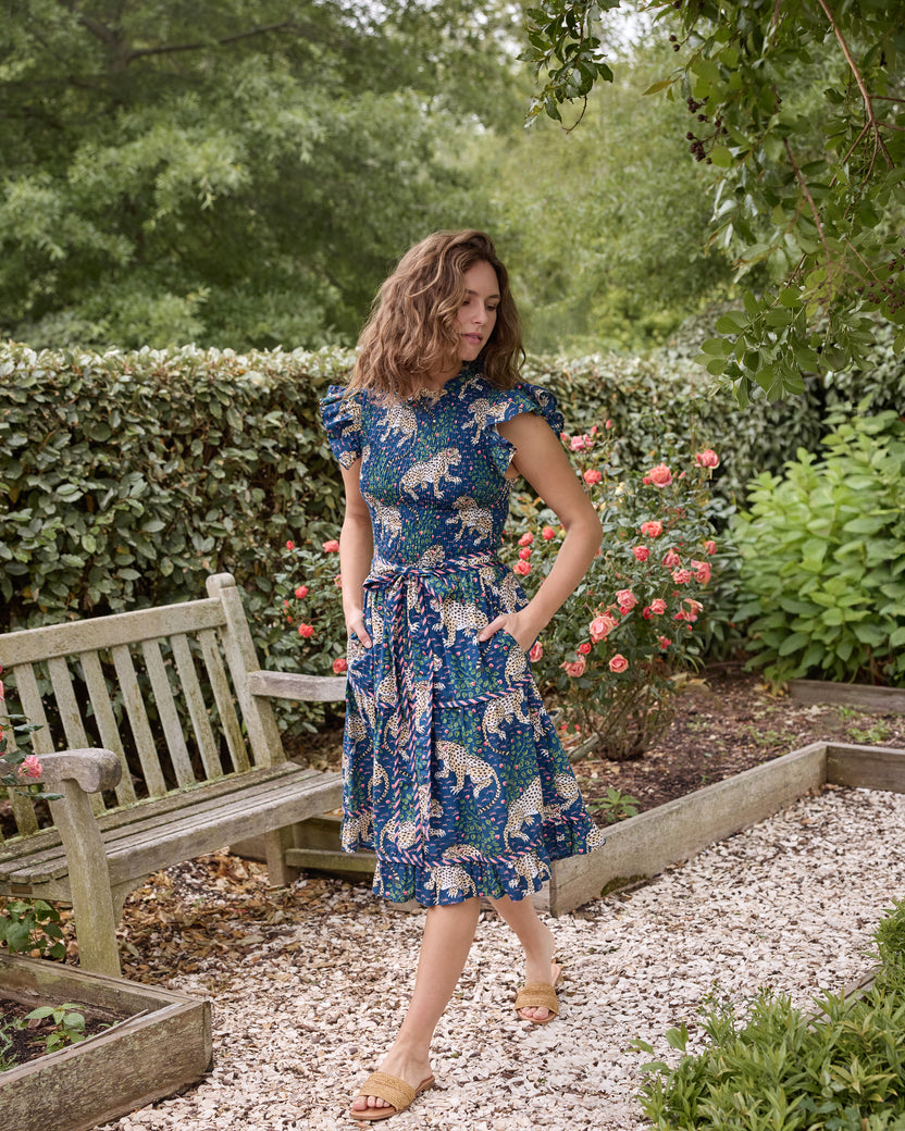 Full-length view of the navy Bagheera Let's Ruffle Dress with leopard print, flutter sleeves, tiered skirt, tied waist belt, and side pockets, worn in a rose garden.