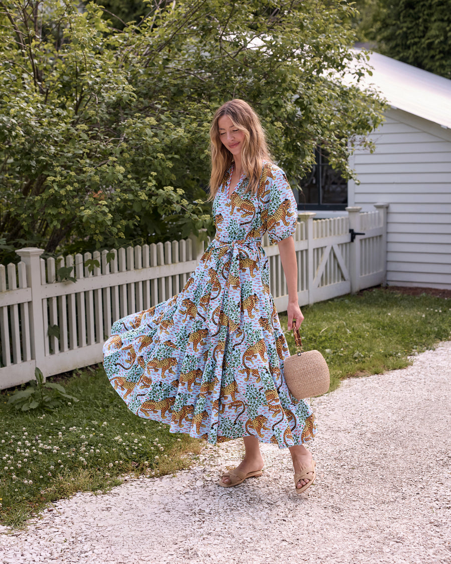 Model wearing the pale blue Bagheera To and From Dress with leopard print, puff sleeves, a tied waist belt, and a full skirt, holding a round woven handbag outdoors.