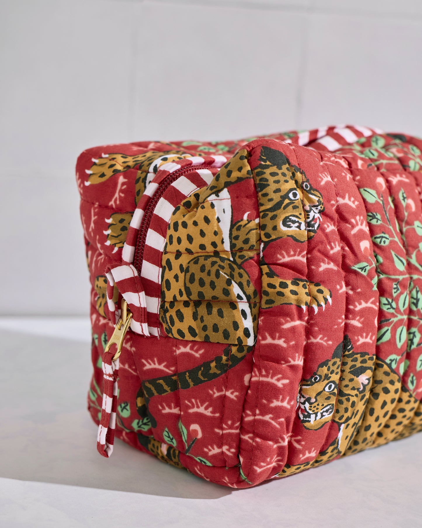 Angled close view of a red quilted Bagheera pouch showing leopard motifs, green leaves, and a red-and-white striped zipper trim.