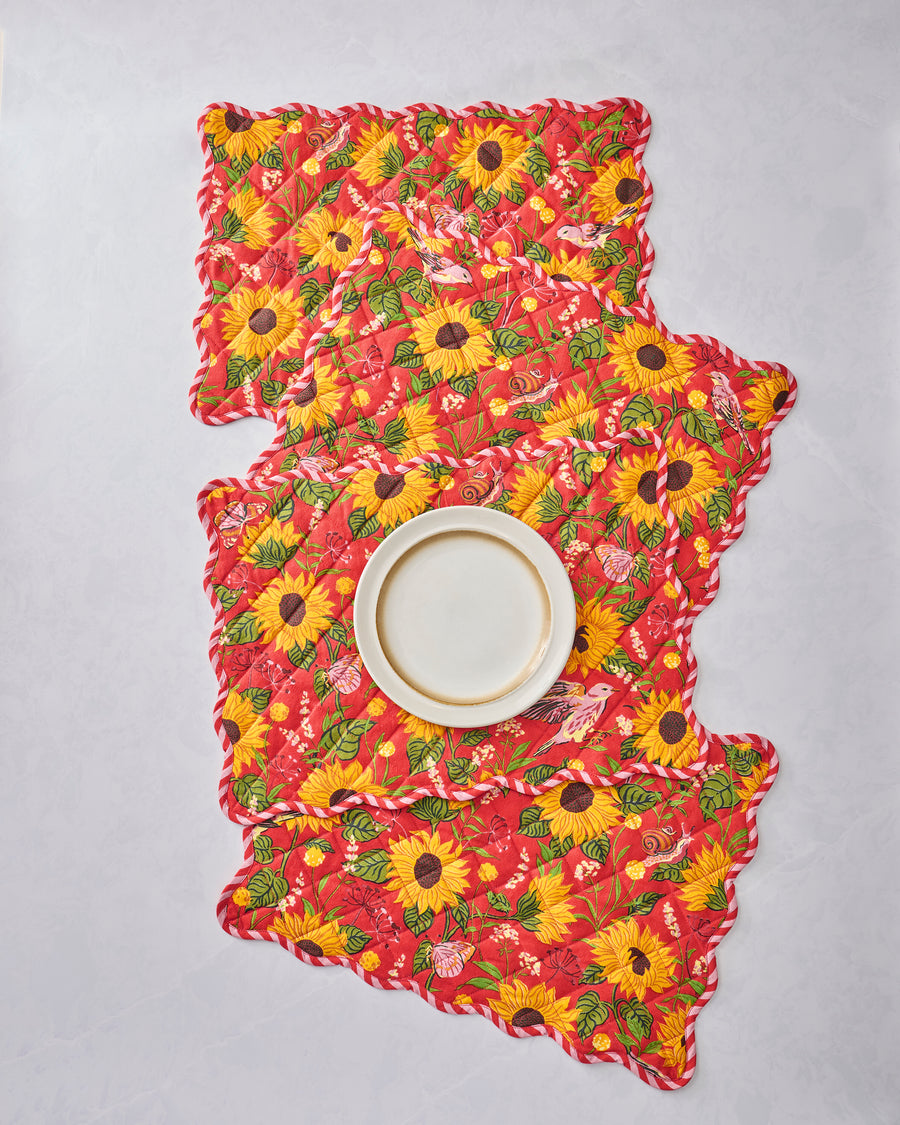 Four orange quilted placemats with scalloped edges and a sunflower, leaf, bird, and butterfly print, arranged beneath a white plate.