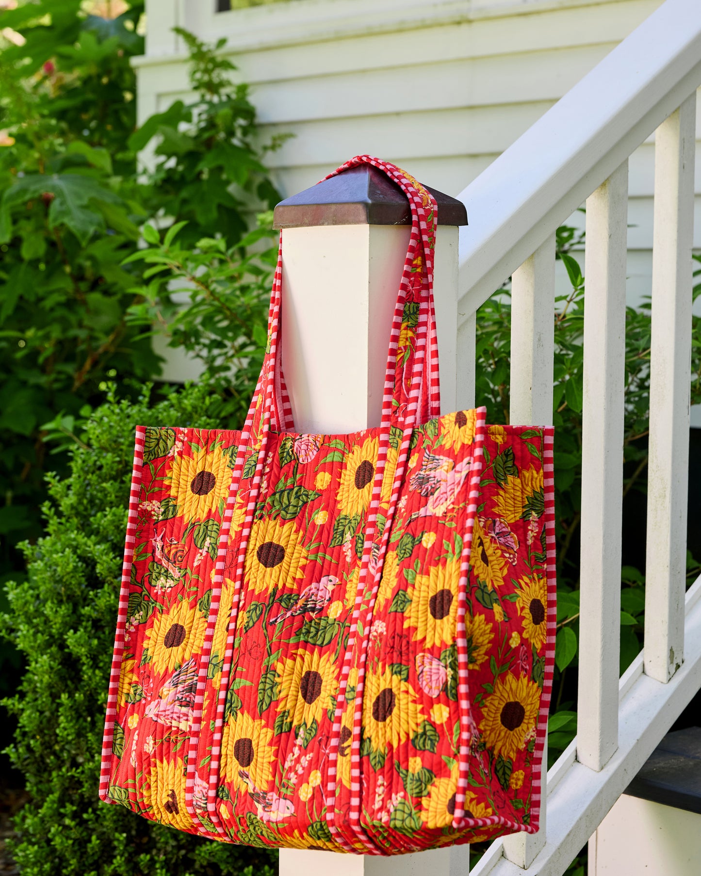 Quilted orange floral tote hanging from a porch post, viewed from the front and side to show its depth and striped handles.