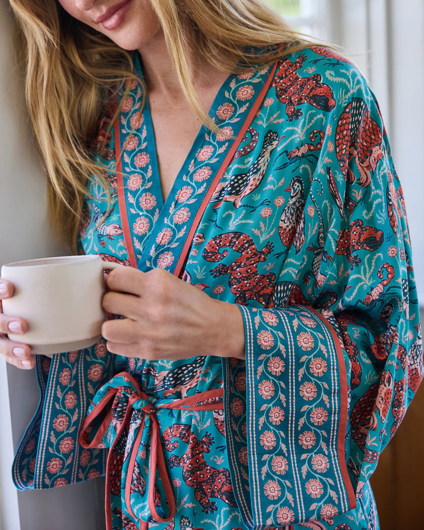 Close-up of the teal satin robe’s wrap front, tie belt, wide sleeve, patterned cuff, and animal print while the wearer holds a cup.