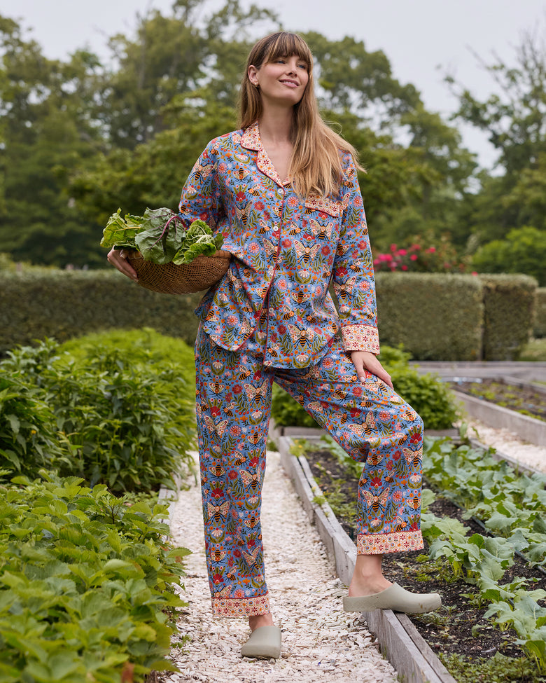 Full view of the Delphinium Blue Bee Tapestry long pajama set in a garden, with the wearer carrying a basket of leafy vegetables.