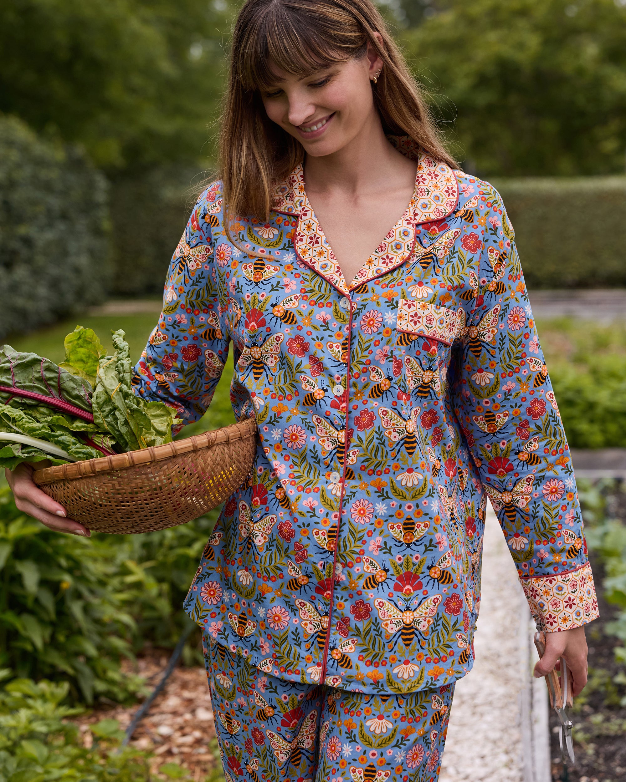 Person wearing the Delphinium Blue Bee Tapestry long pajama set and carrying a woven basket of leafy vegetables in a garden.