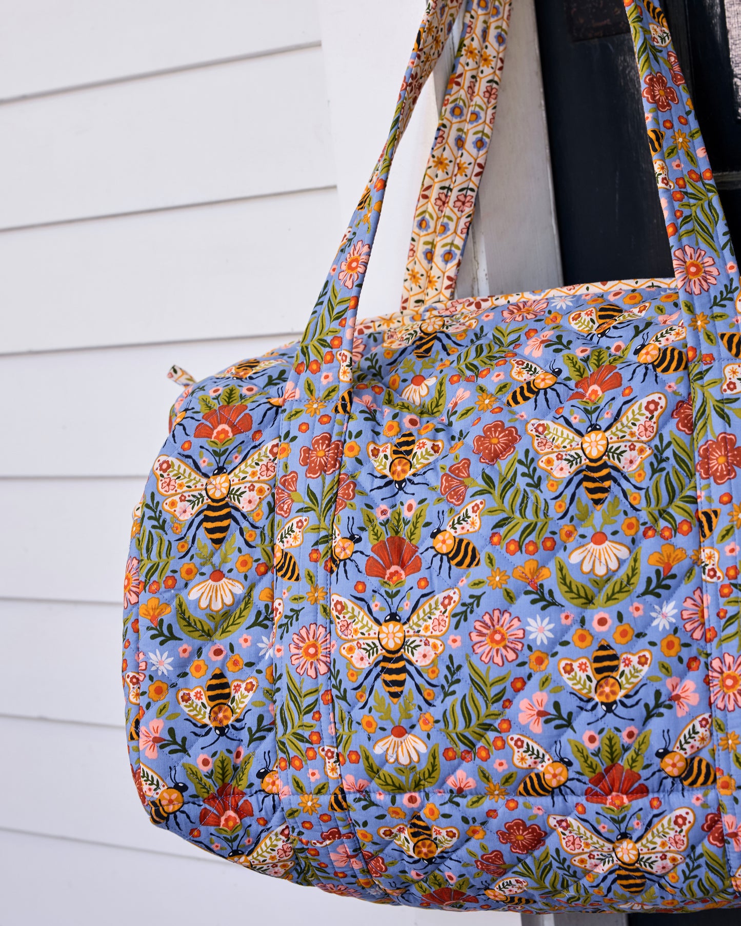 Close-up of the Bee Tapestry quilted duffle bag showing its blue bee-and-floral print, quilted seams, and handles.