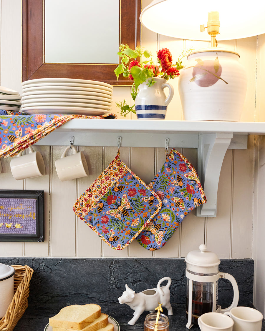 Blue floral oven mitt and matching square pot holder hanging in a kitchen beside cups and dishes.