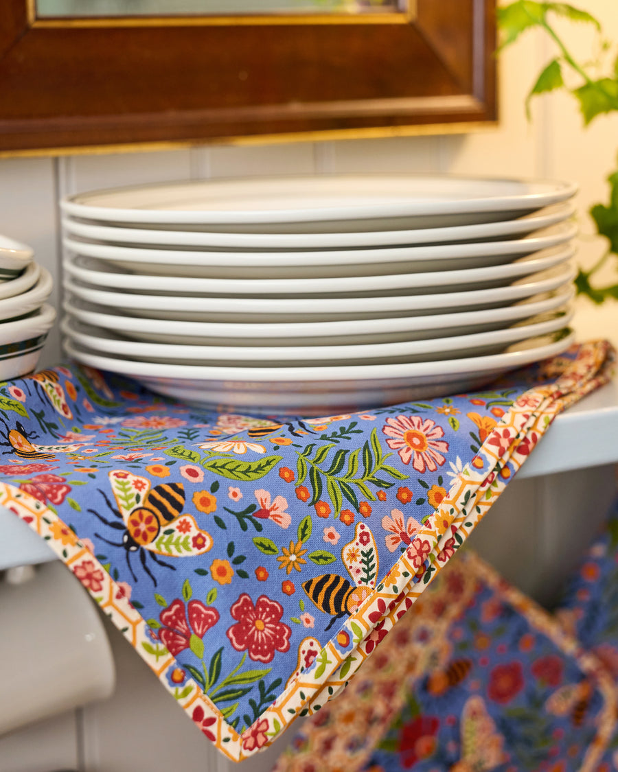 Blue Bee Tapestry tea towel draped beneath a stack of white plates, with bees, flowers, leaves, and a floral patterned border.