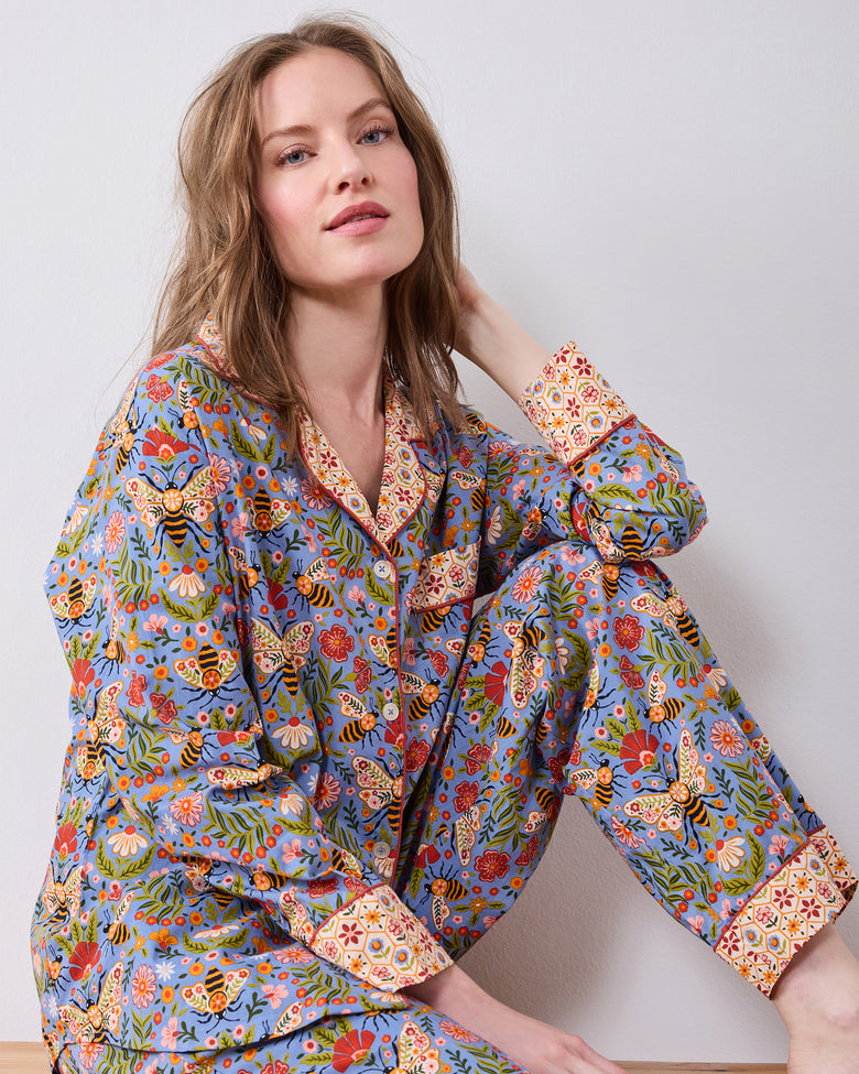 Seated model wearing a long-sleeve button-front pajama top and matching pants with a blue bee-and-floral print and contrasting patterned collar and cuffs.