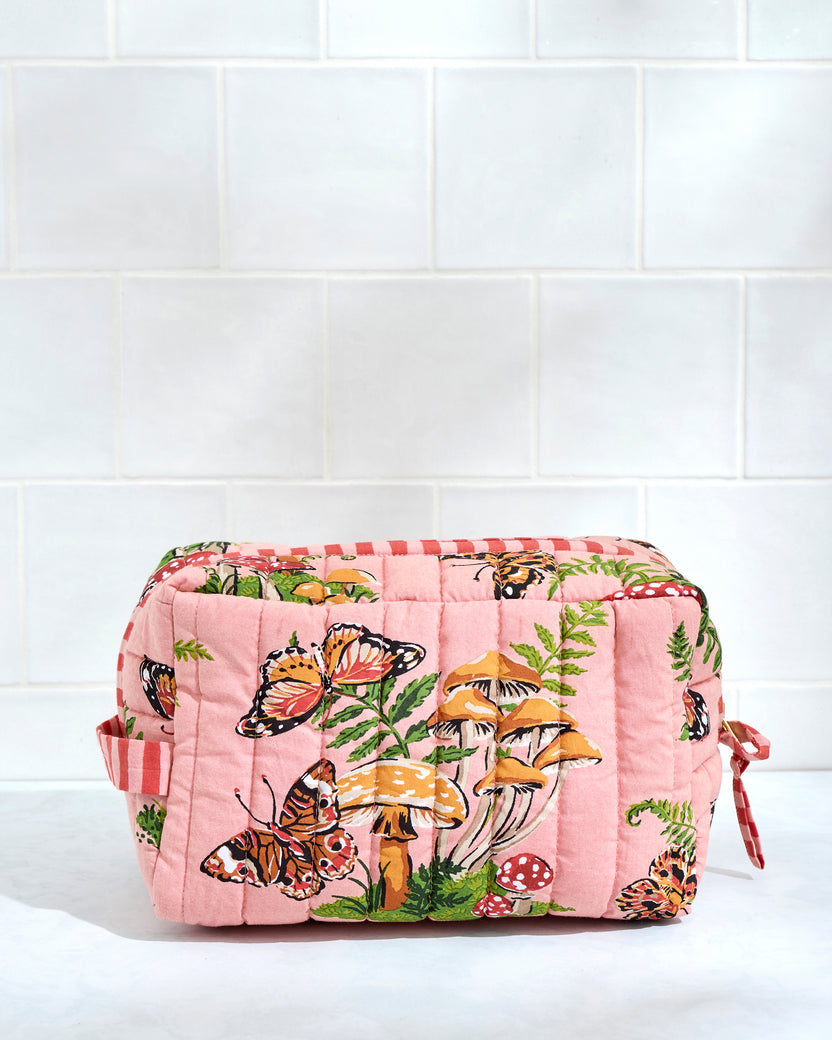 Front view of a quilted butterfly-print pouch with mushrooms and ferns, positioned against a white tile wall.