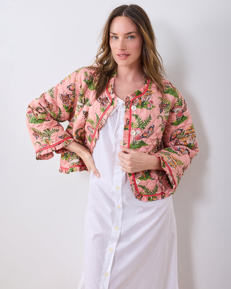 Front view of a person wearing the quilted pink Butterfly Forager slumber jacket over a white button-front top, showing the full jacket, ruffled cuffs, trim, and woodland print.