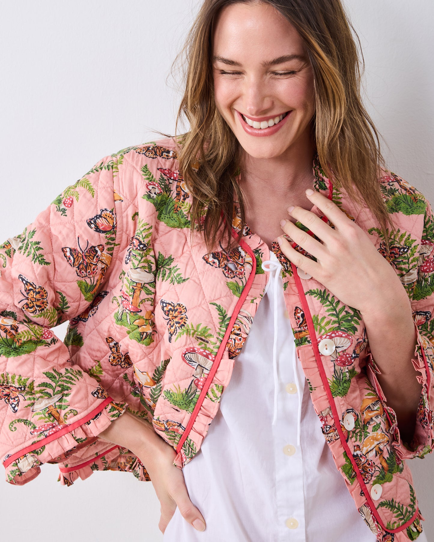 Front three-quarter view of a person wearing the quilted pink Butterfly Forager slumber jacket over a white top, showing ruffle trim, buttons, and the butterfly-and-mushroom print.