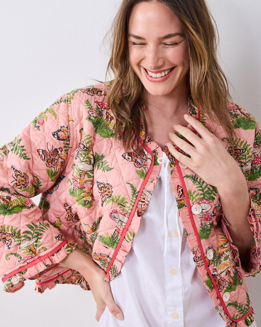 Front three-quarter view of a person wearing the quilted pink Butterfly Forager slumber jacket over a white top, showing ruffle trim, buttons, and the butterfly-and-mushroom print.