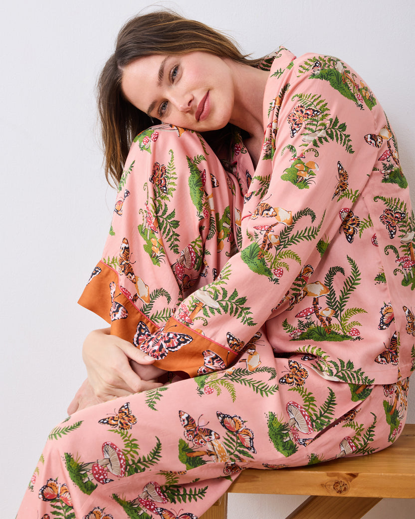 Close view of a model seated on a wooden bench wearing the pink Butterfly Forager tall pajama set, showing the patterned top, pants, and contrasting cuffs.