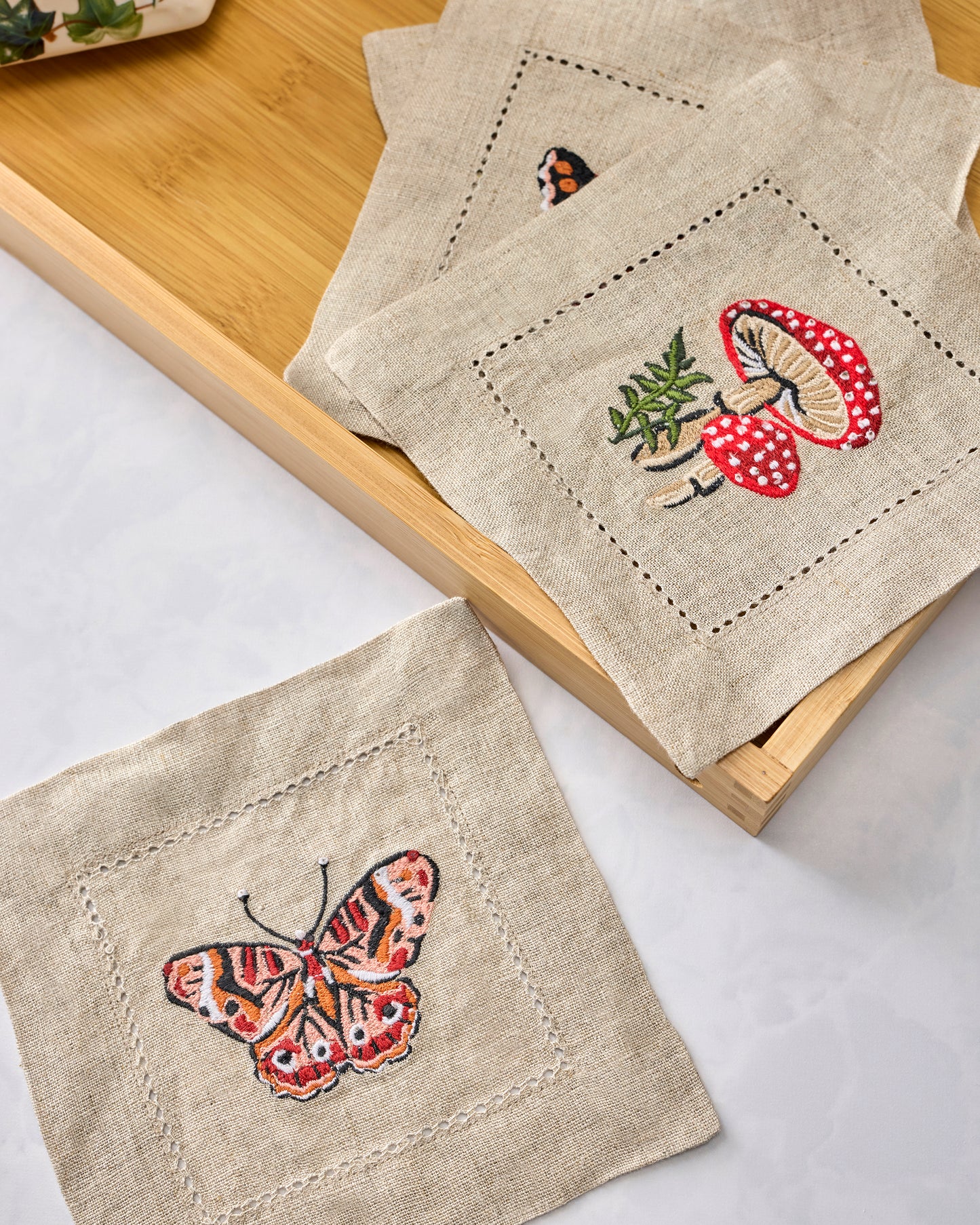 Four embroidered cocktail napkins arranged on a wooden tray, featuring butterfly, mushroom, and botanical designs.