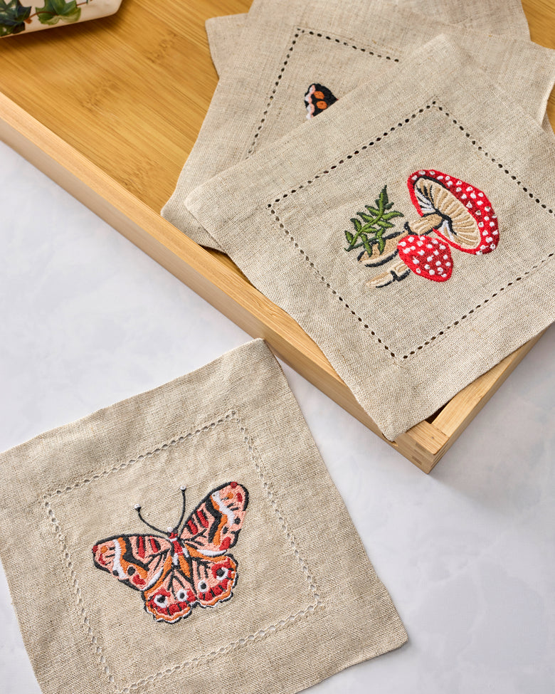 Four embroidered cocktail napkins arranged on a wooden tray, featuring butterfly, mushroom, and botanical designs.