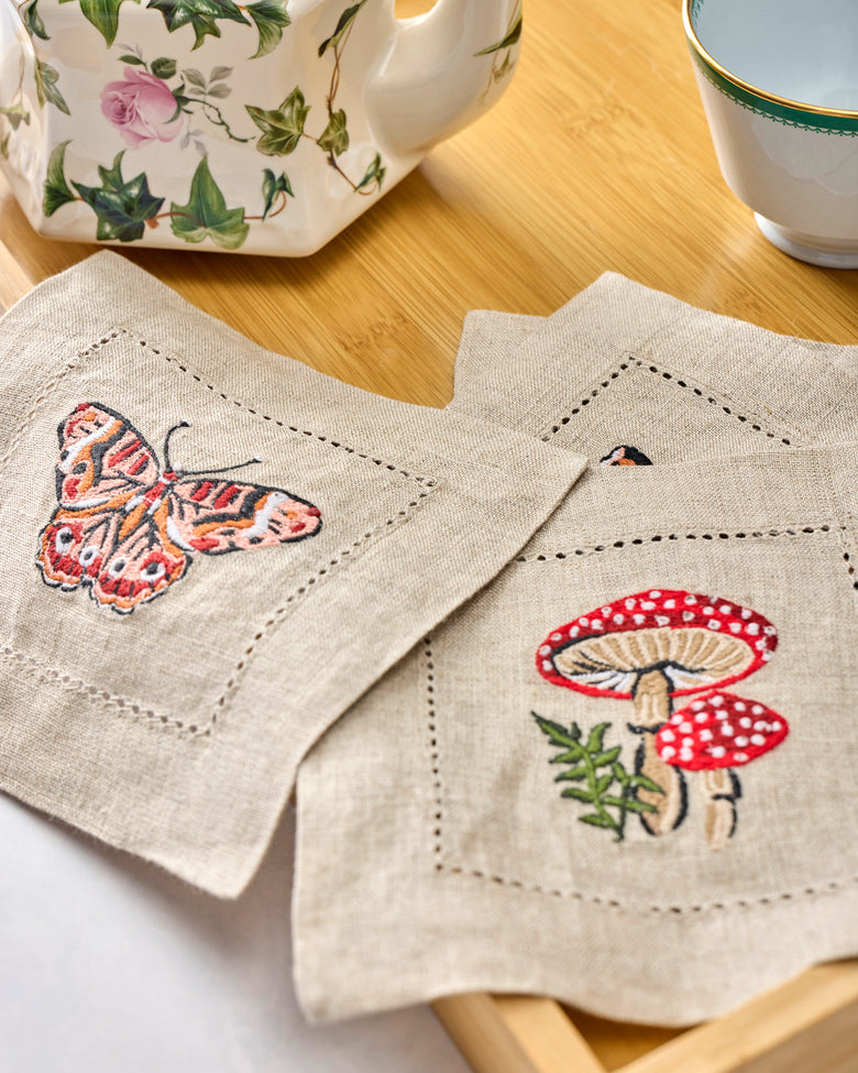 Close view of four embroidered cocktail napkins with butterfly and mushroom designs arranged on a table.