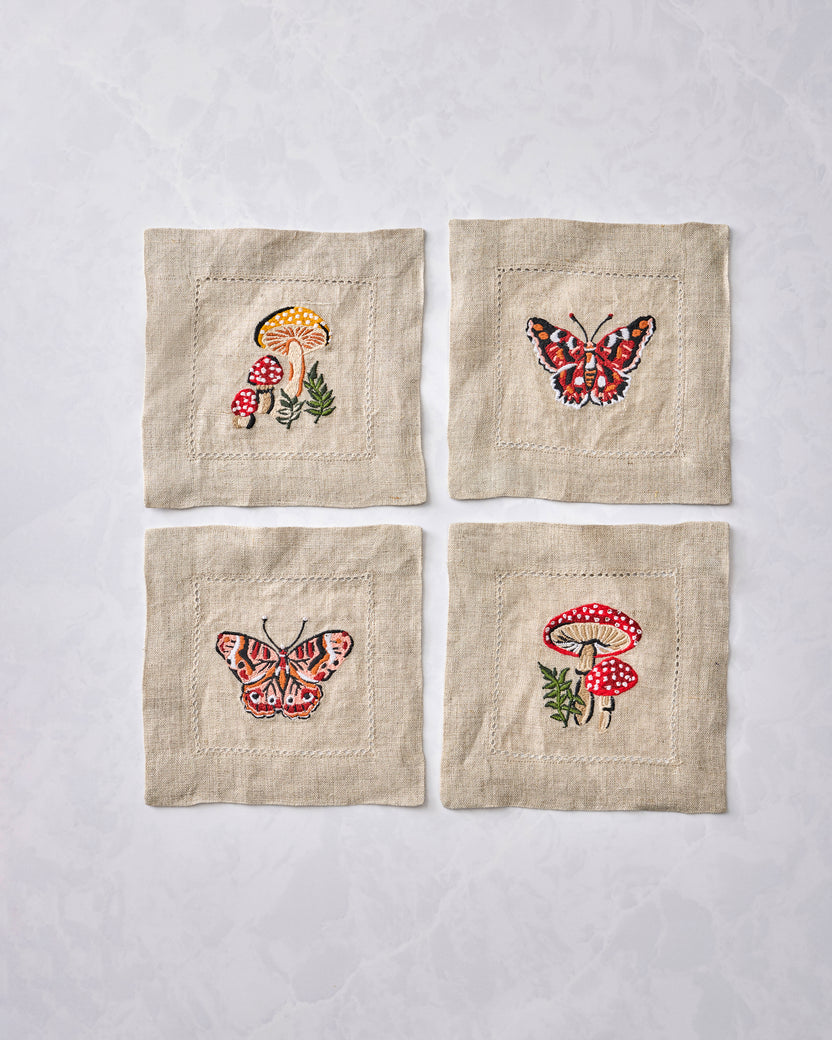 Four square embroidered cocktail napkins displayed in a two-by-two arrangement, with mushroom and butterfly designs.