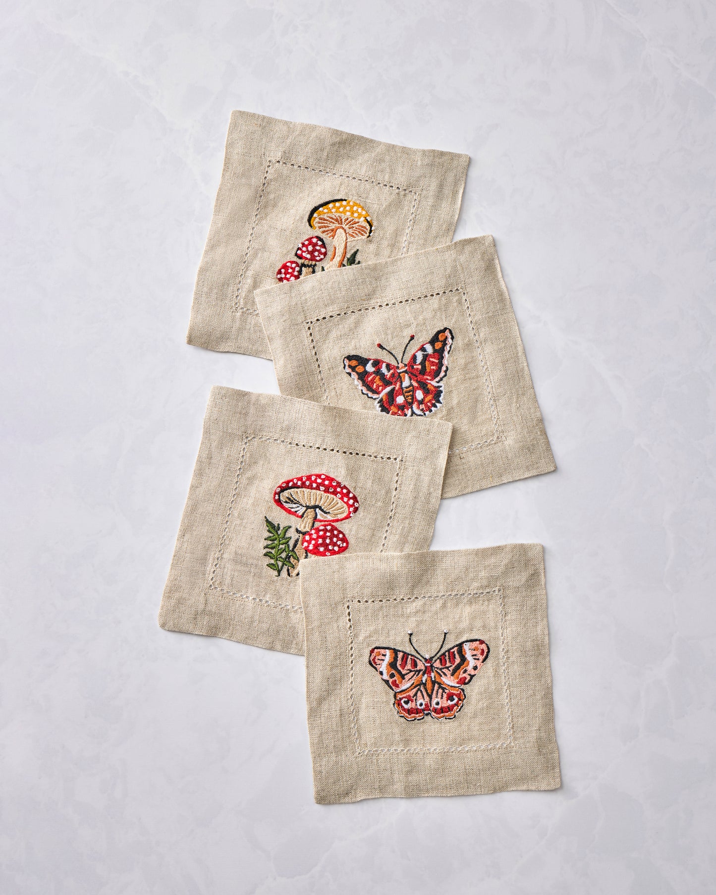 Four embroidered cocktail napkins overlapping in a diagonal arrangement, showing mushroom and butterfly designs.
