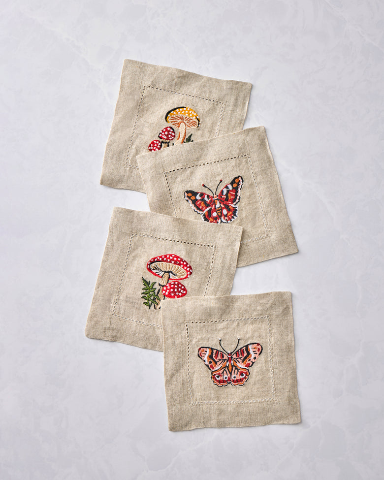 Four embroidered cocktail napkins overlapping in a diagonal arrangement, showing mushroom and butterfly designs.