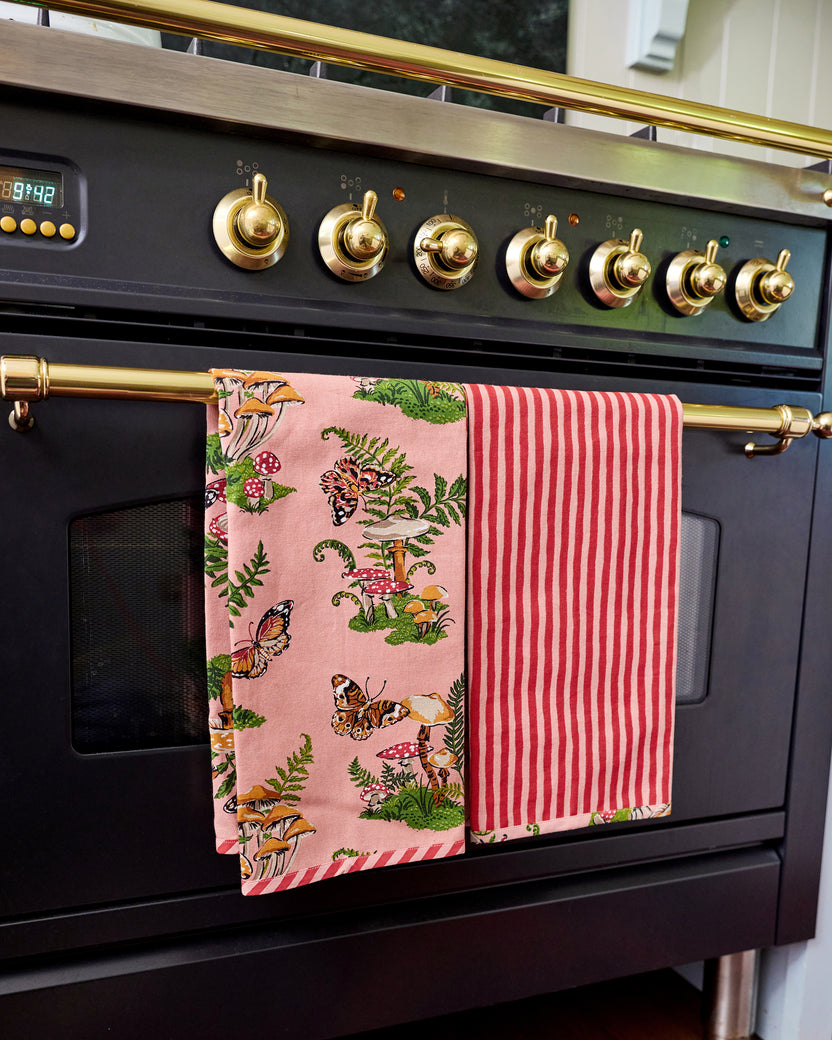 Two Butterfly Forager tea towels hanging from an oven handle, one showing the pink woodland print and the other showing red-and-white vertical stripes.