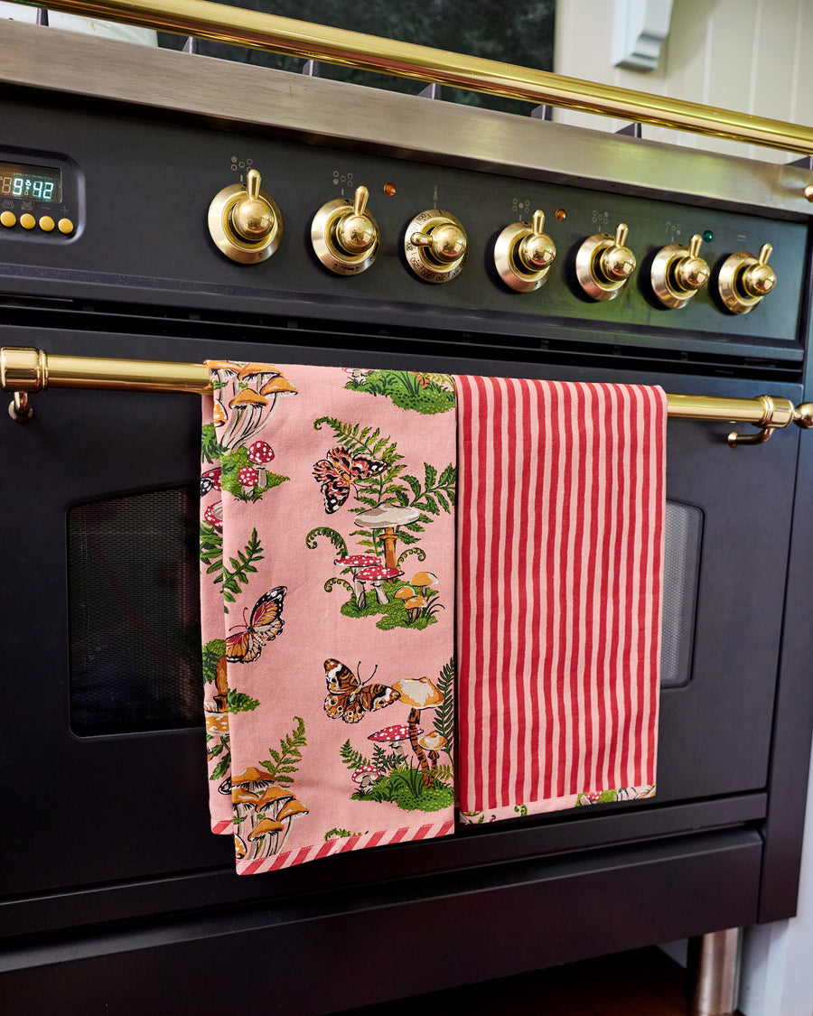 Two Butterfly Forager tea towels hanging from an oven handle, one showing the pink woodland print and the other showing red-and-white vertical stripes.