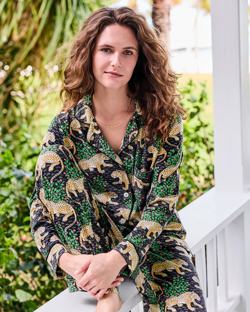 Person seated on a porch railing wearing a black long-sleeve pajama set with a leopard-and-floral print.
