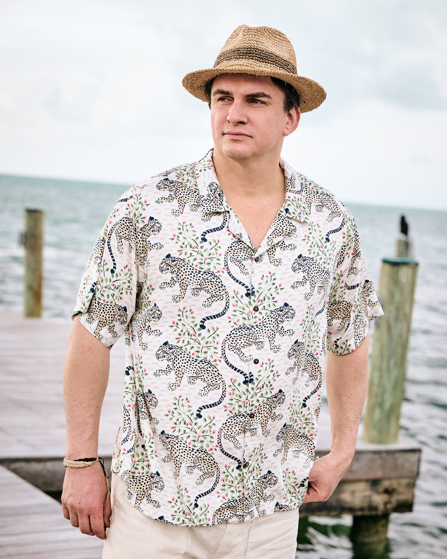 Model wearing the Wedding Cake Bagheera short-sleeve camp shirt with light trousers and a straw hat by the water