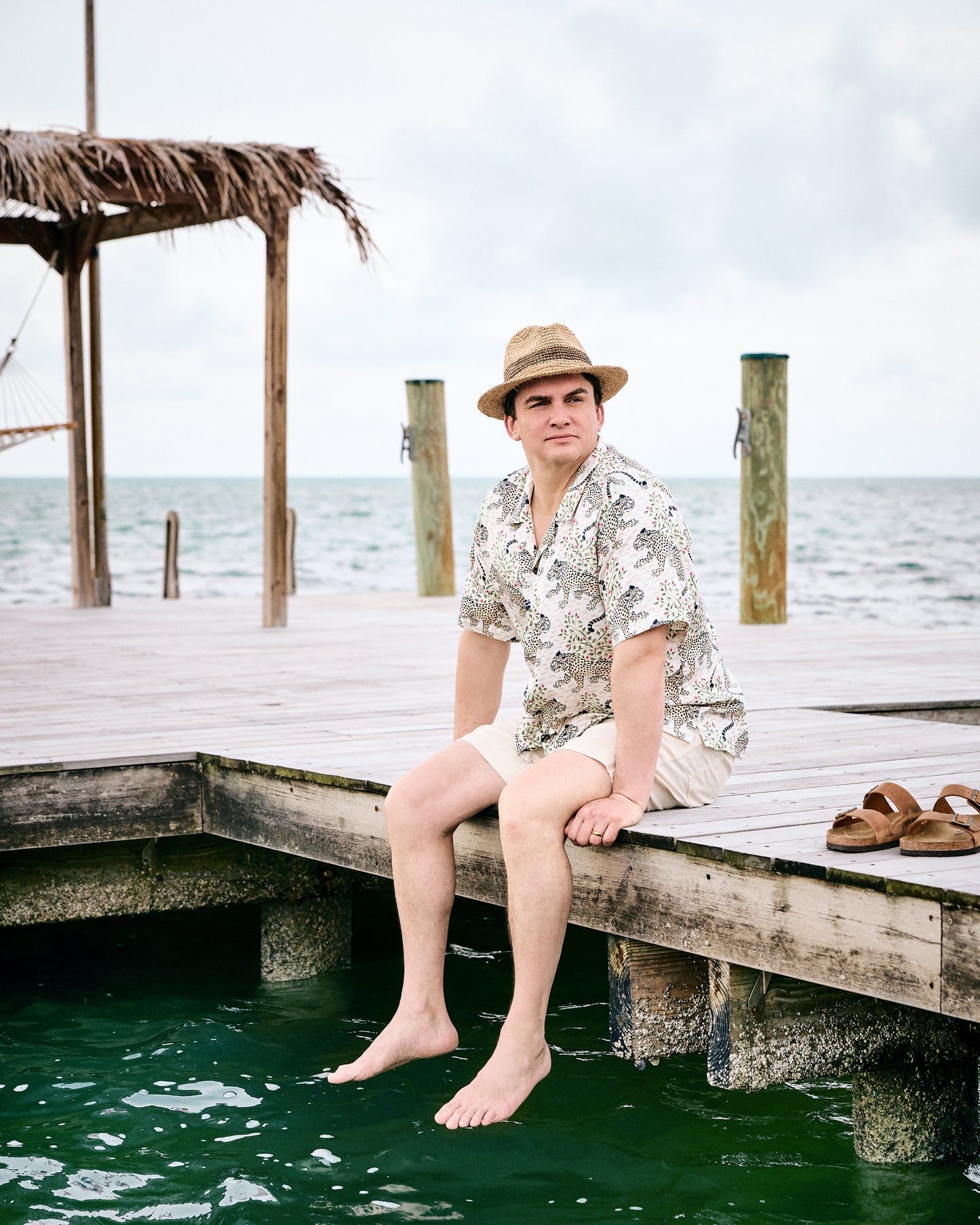 Model seated on a dock wearing the Wedding Cake Bagheera camp shirt, straw hat, and shorts