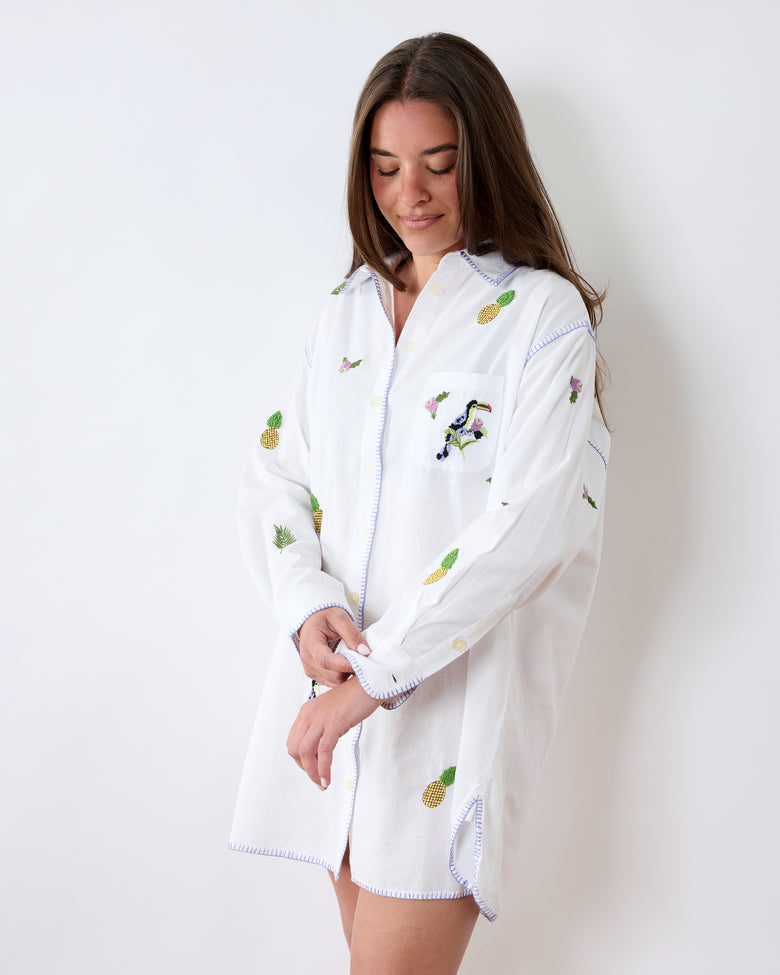 Angled front view of a white oversized button-down sleep shirt with pineapple, leaf, flower, and toucan embroidery.