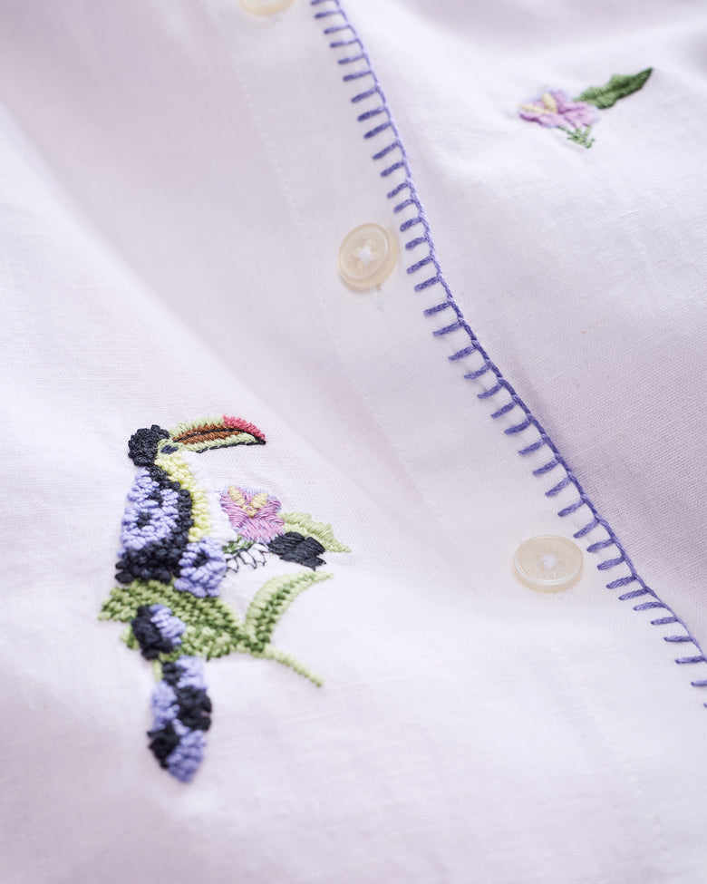 Close-up of the white button-down showing pineapple-and-toucan embroidery, cream buttons, and purple stitching along the placket.