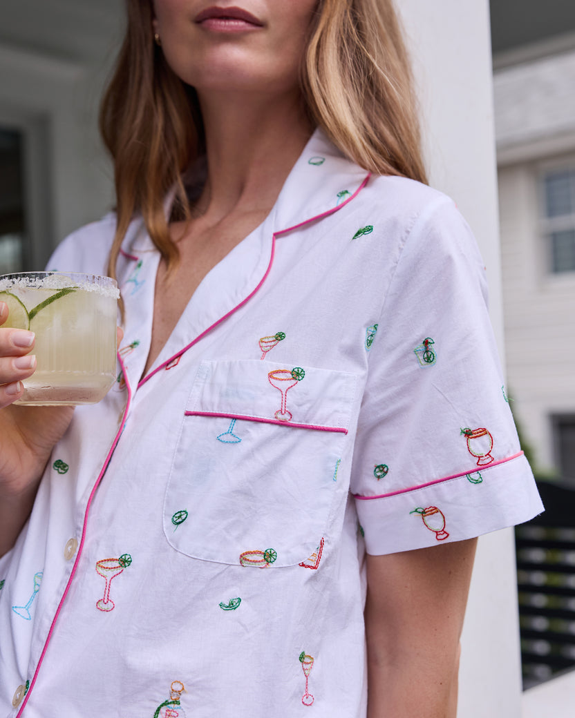 Close-up of the cloud pajama top showing pink piping, a chest pocket, buttons, and embroidered cocktail-themed motifs; a glass is partly visible.