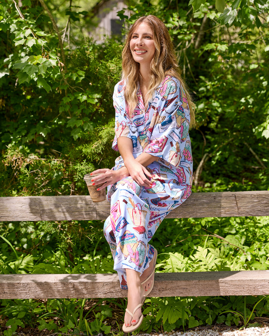 Person wearing a pale blue short-sleeve button-front pajama set with a travel-themed print, seated on a wooden fence and holding a takeaway cup.