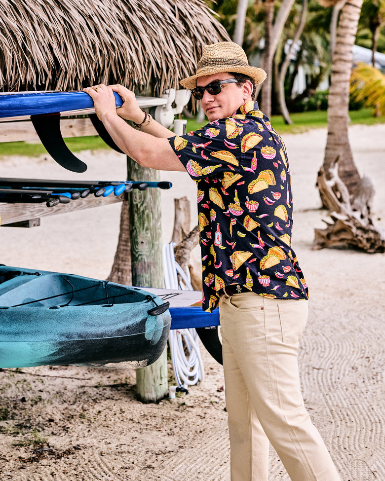 Model wearing the dark short-sleeve shirt with an allover taco, nacho, pepper, and hot-sauce print beside a kayak rack
