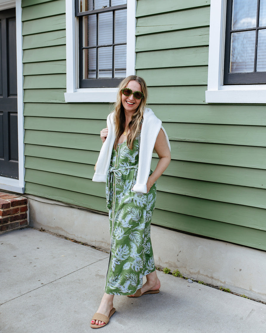 Person wearing the green Heron Landing Alma dress with a white sweater draped over the shoulders and sunglasses outdoors.