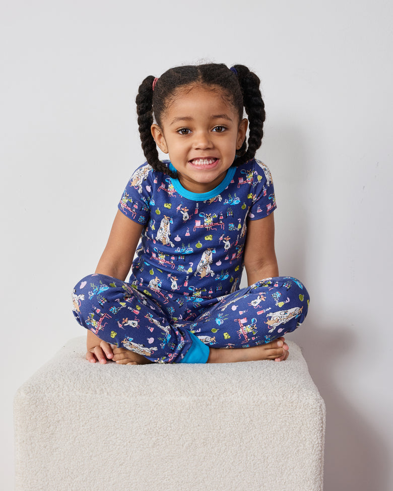 Child seated cross-legged wearing a blue short-sleeve top and pants set with a colorful laboratory-themed print.