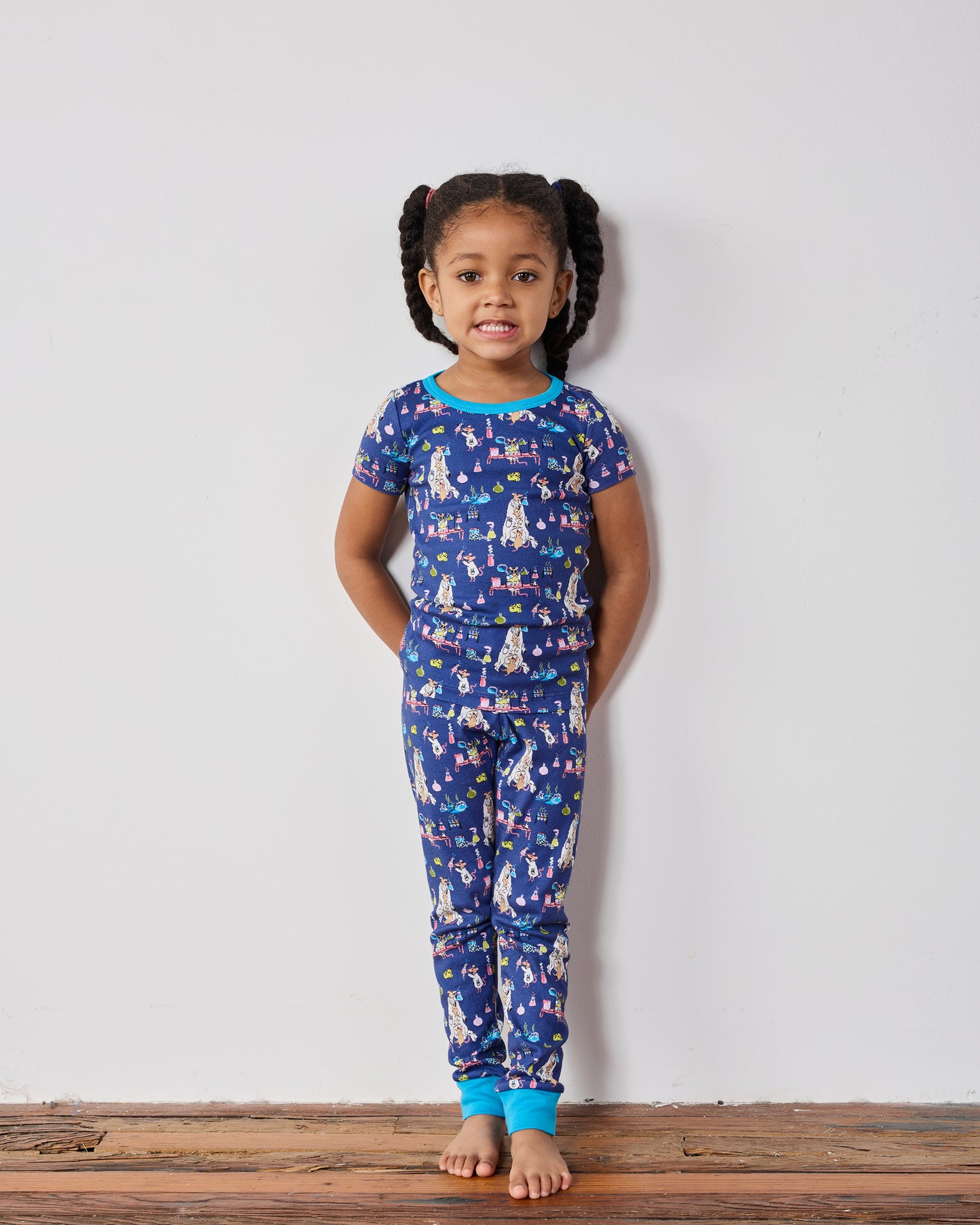Child standing against a wall wearing the complete blue short-sleeve pajama set with a colorful laboratory-themed print.
