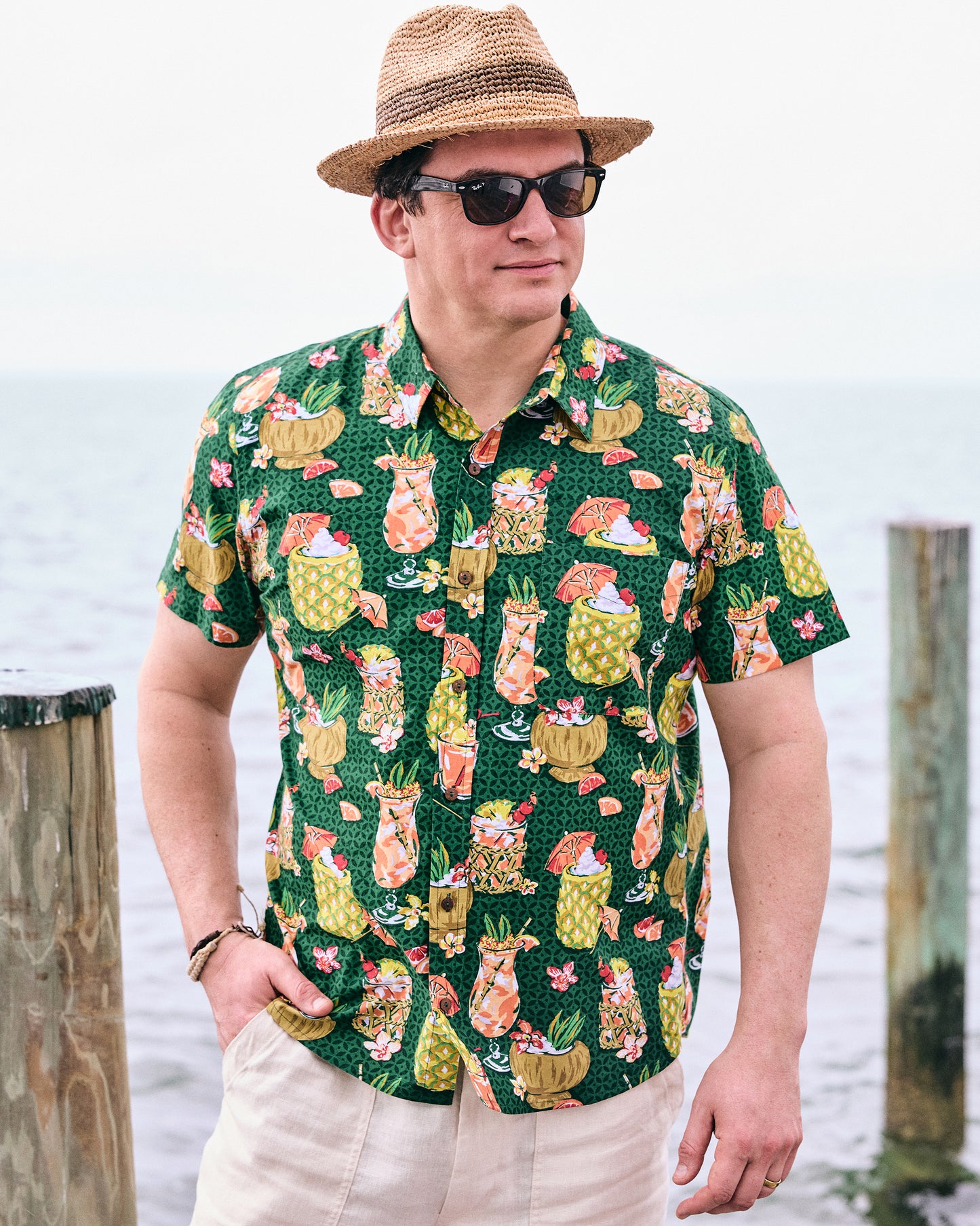 Model wearing the emerald green tropical drink-print shirt with beige trousers, a straw hat, and sunglasses by the water