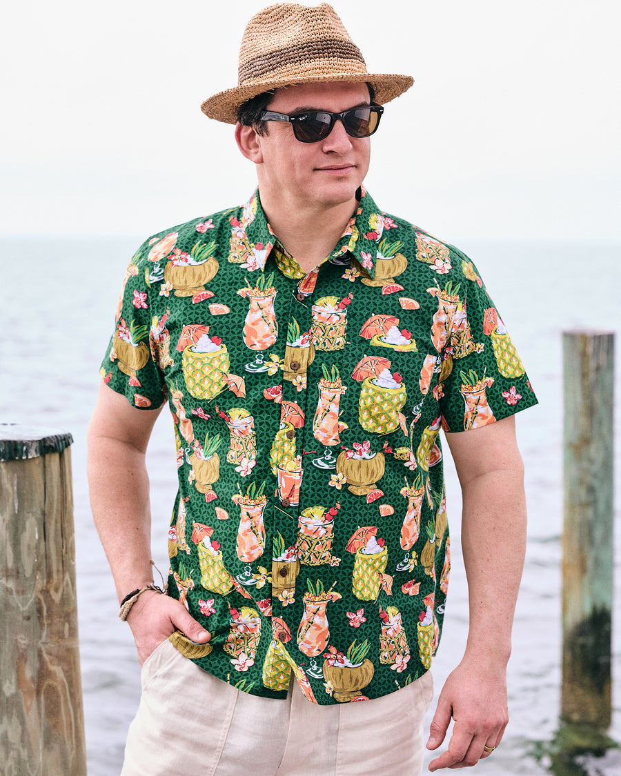Model wearing the emerald green tropical drink-print shirt with beige trousers, a straw hat, and sunglasses by the water