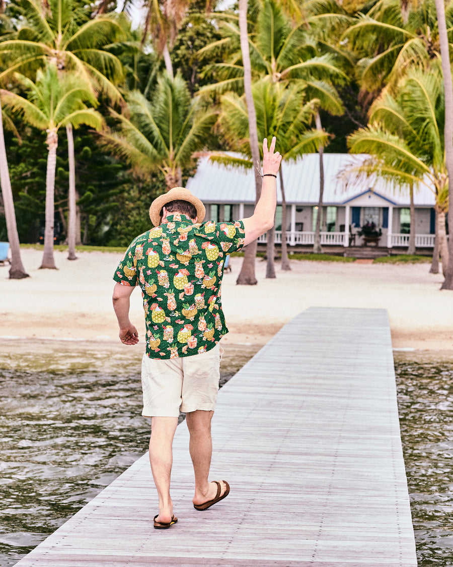 Back view of the emerald green tropical drink-print shirt worn on a dock lined with palm trees