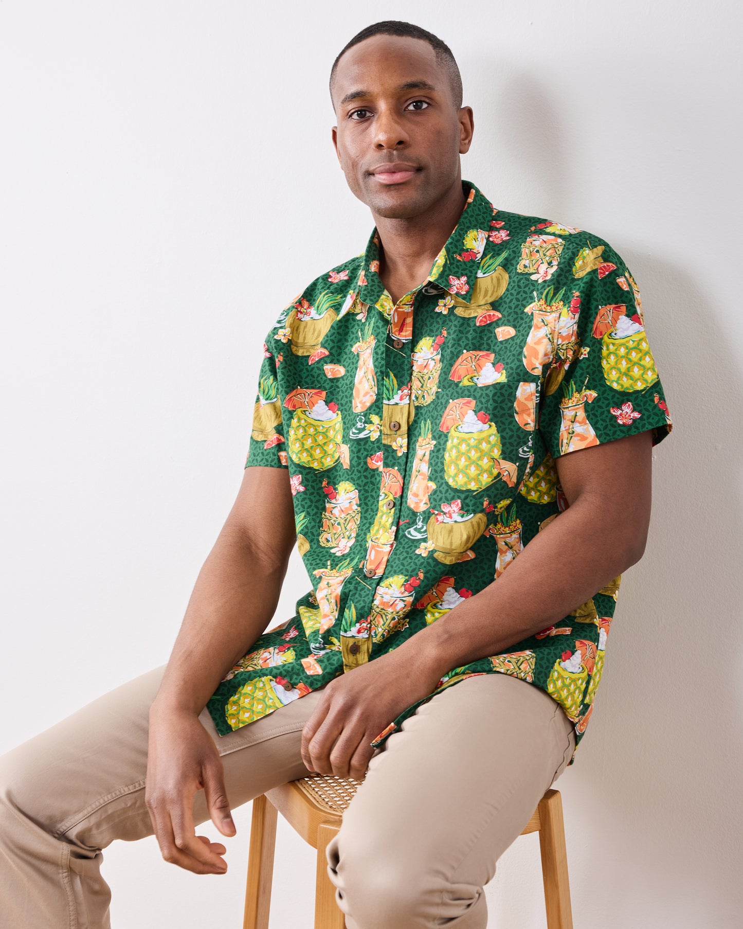 Seated model wearing the emerald green tropical drink-print shirt with beige trousers