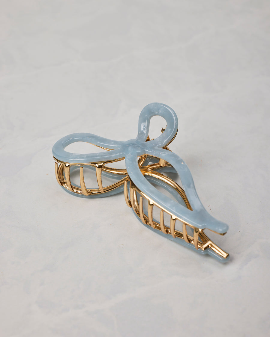 Pale blue ballet bow claw hair clip shown at an angle, with gold-toned teeth and frame.