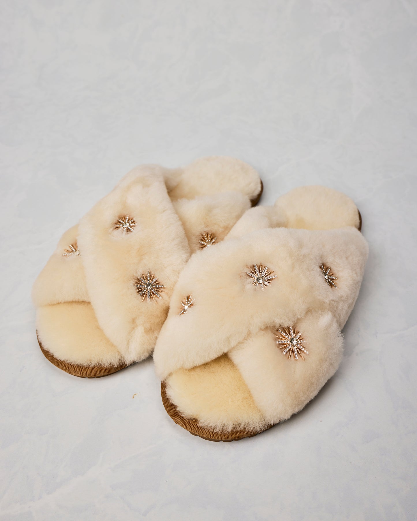 Pair of beige sheepskin slippers with gold-toned rhinestone starburst embellishments.