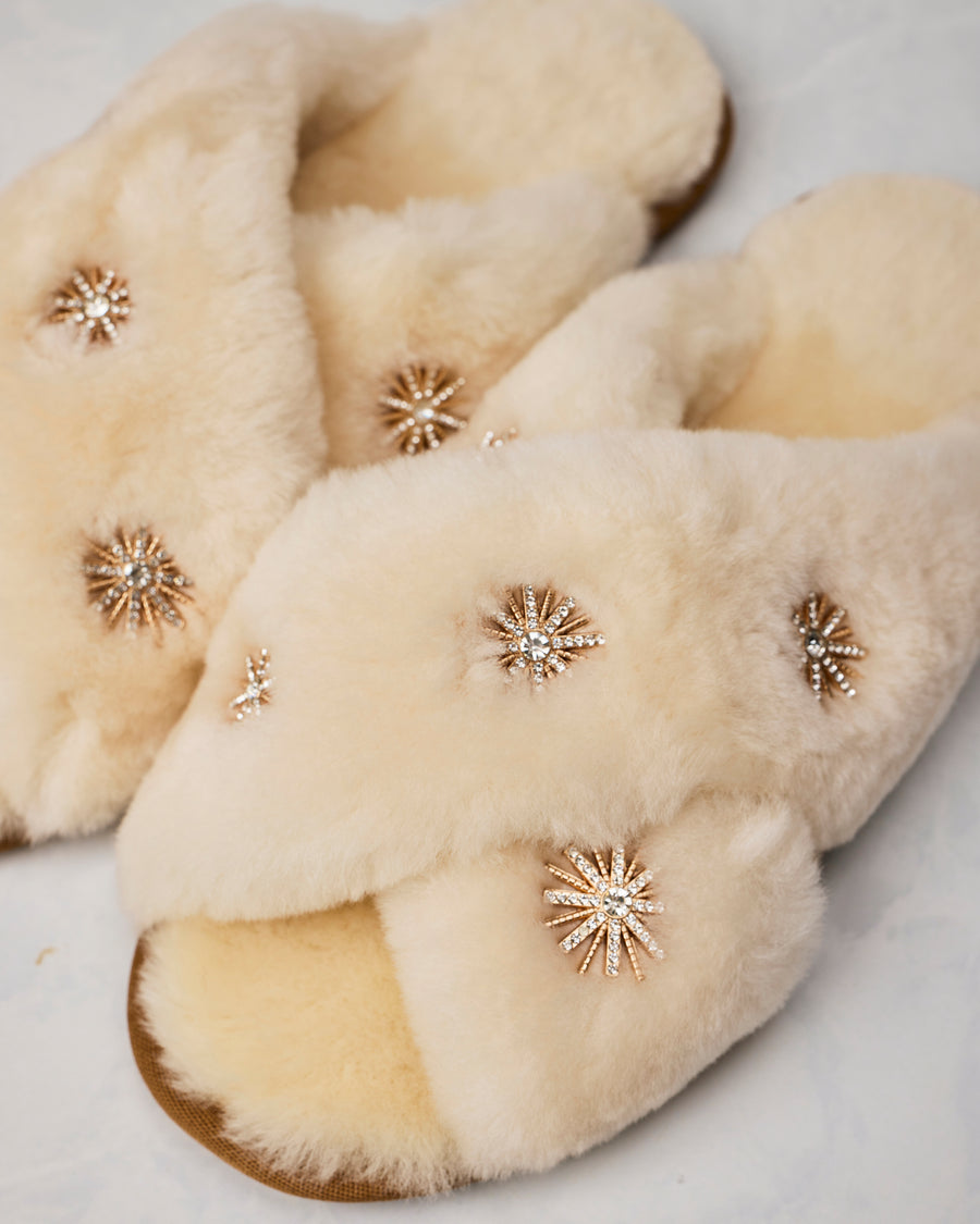 Close-up of beige sheepskin slippers showing the fuzzy crossed uppers and rhinestone starburst embellishments.