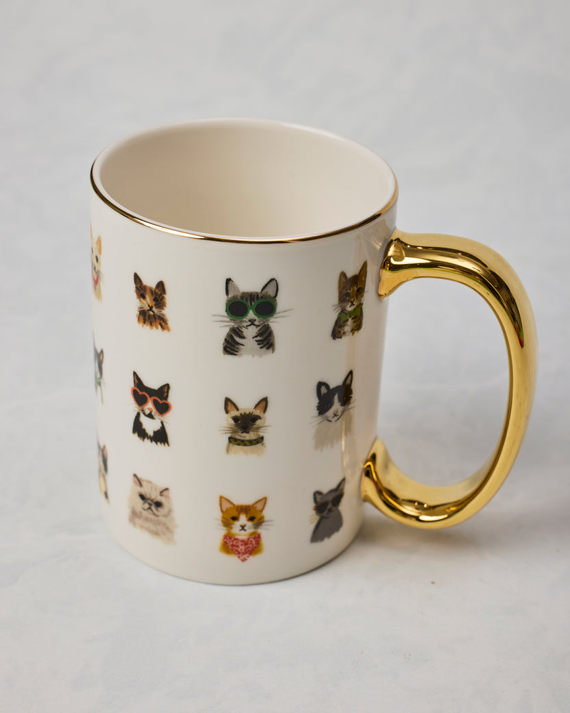 White mug with illustrated cats wearing accessories and a gold-colored handle and rim